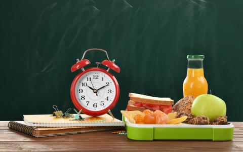 A photograph of an alarm clock surrounded by a school lunch tray and school supplies.