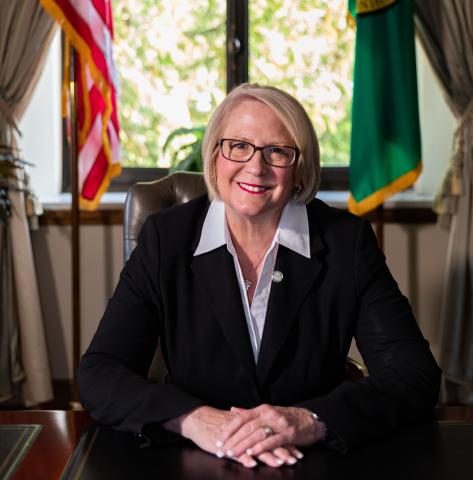 State Auditor Pat McCarthy sitting at desk with a U.S. and Washington state flag behind her