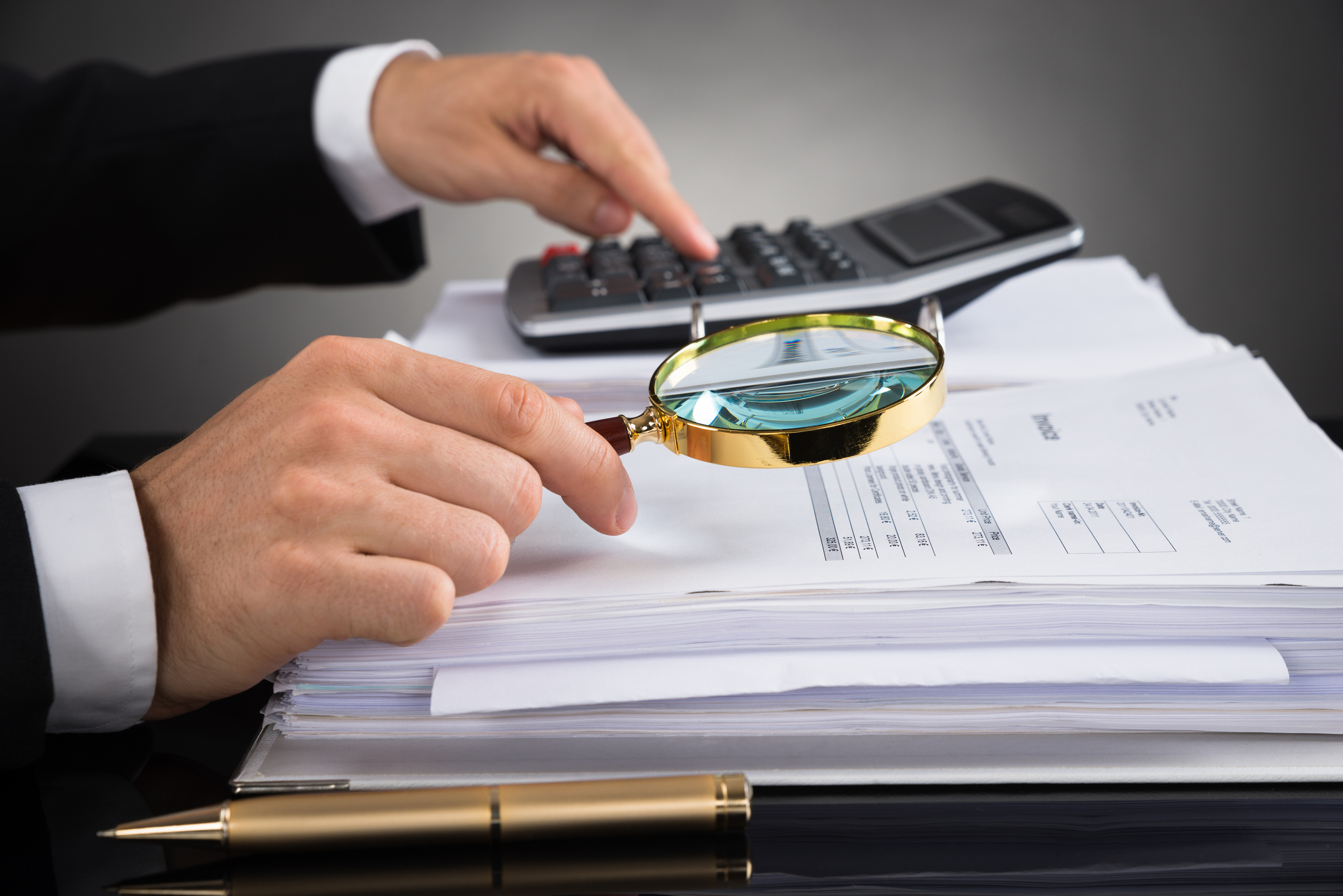 Close-up Of Businessperson Hands Checking Invoice With Magnifying Glass At Desk