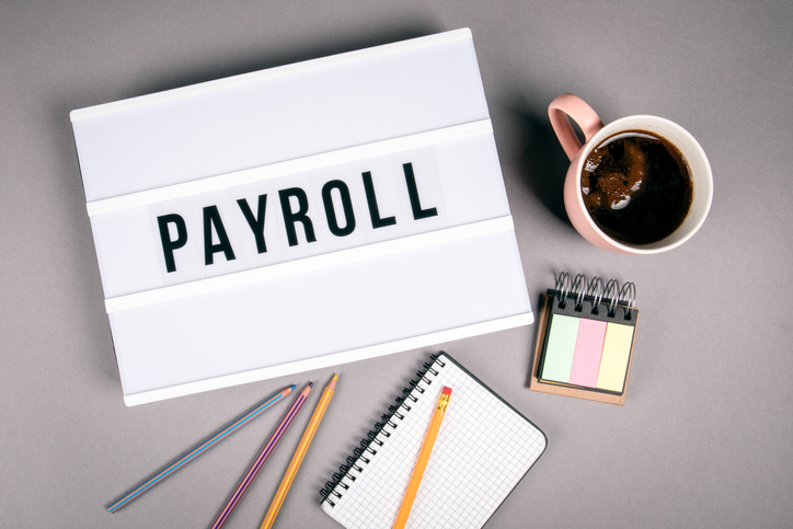 Concept image of payroll. There's a white binder with the word payroll written on it. The binder is surrounded by a mug of coffee, four pencils, a notepad, and pad of sticky notes.